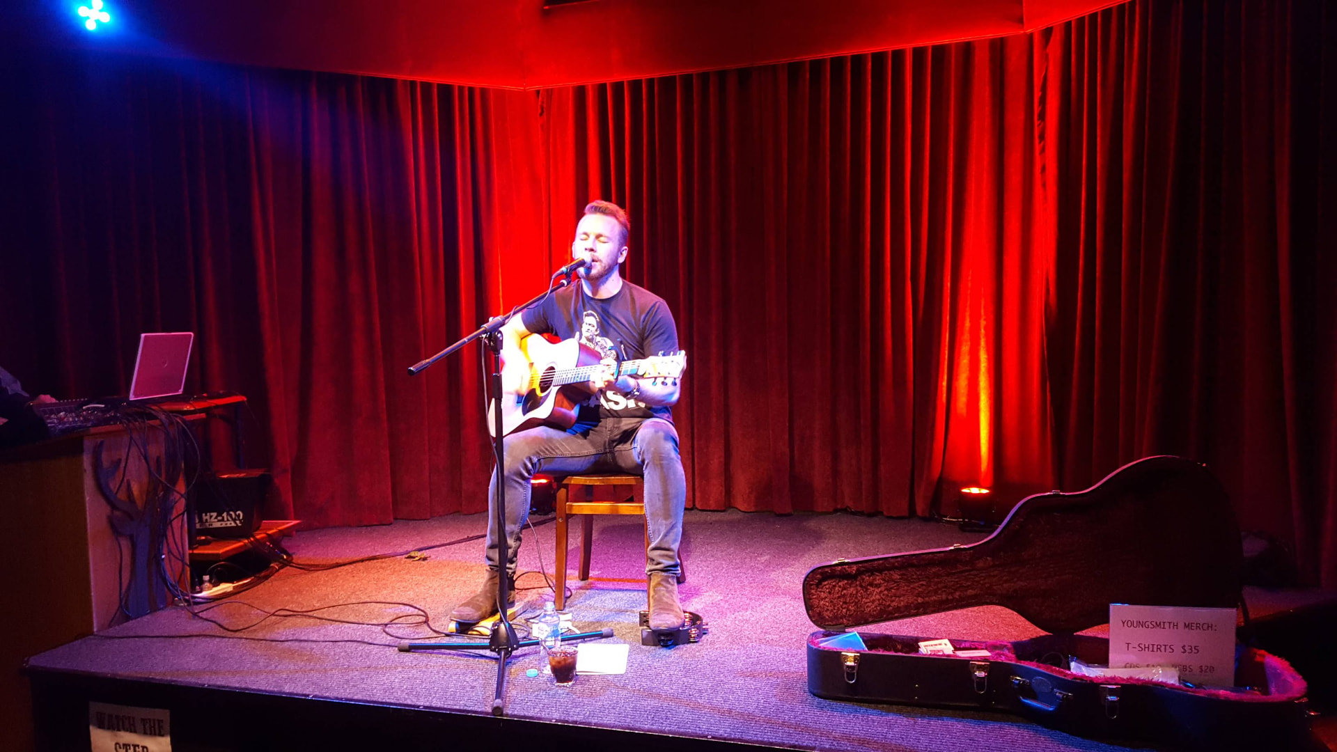 Dan Smith performing alone on a red curtain stage with guitar case open beside him